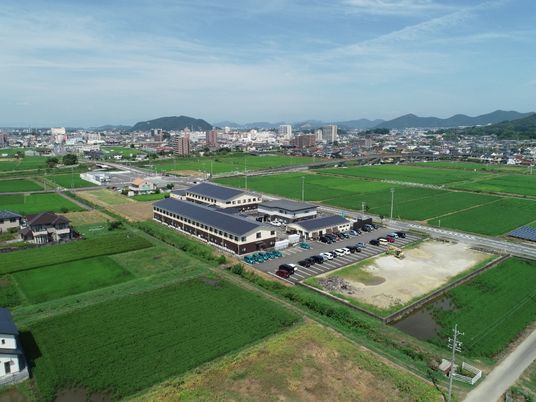 外壁が白とブラウン、屋根がグレーの２階建ての建物が複数同じ敷地内にあり、周辺には緑の田畑が広がり、奥には住宅地がある。