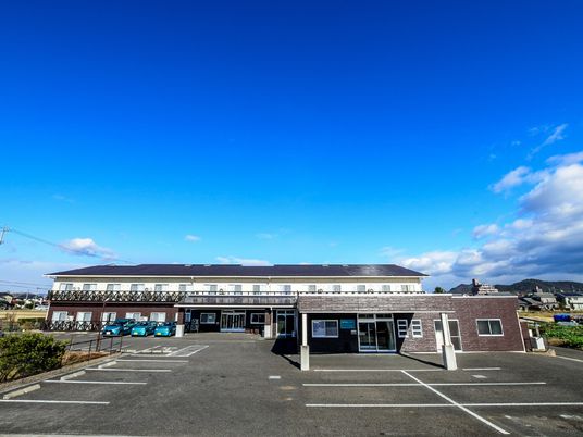 青い空の下に建っている2階建ての建物。1階部分がブラウンで、2階部分が白、広い駐車場があり、周りには畑も広がっている。