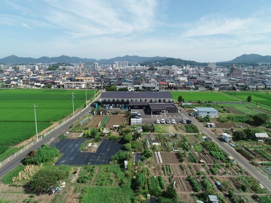 田畑に囲まれた白とグレーの2階建ての建物は和の雰囲気が漂う。奥には住宅地が広がっていて、山脈も見ることができる。