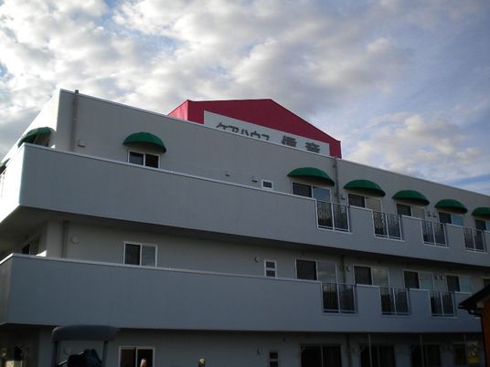 ３階建ての建物の各フロアにベランダがあり、一番上には赤い大きな看板のようなものがあり、施設名が書かれている。
