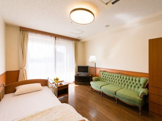 ベッドの横にソファが置いてあるワンルームタイプの部屋の様子。やわらかい照明、窓の外にベランダ、手前に収納設備