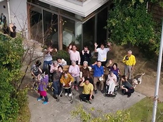 庭に集まり上空を見上げて写真撮影に応じている入居者とスタッフたち。車いすや歩行器の利用者もいる。