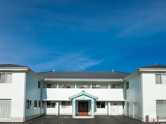 2階建ての建物は白い外壁でグレーの屋根が付き、2階部分にはベランダもある。