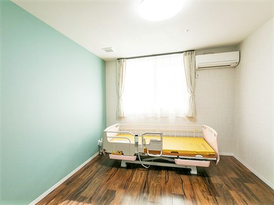介護ベッドが置かれたフローリングの居室には大きな窓が付き、白い壁の一面だけミントグリーン。