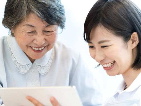 カルテのようなものを見ながら笑顔で話をしている若い女性スタッフとレースの襟が付いたブラウスを着た高齢女性。