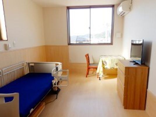 介護ベッドやデスク、チェスト、テレビなどが置かれたフローリングの部屋。