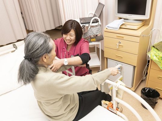 聴診器を肩にかけた看護師がベッドに座った女性に対して笑顔で対応している。