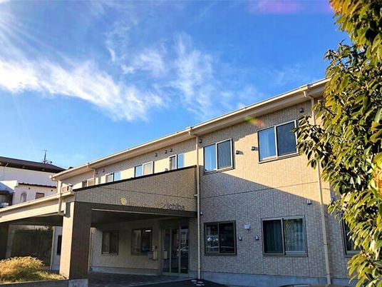 日当たりのいい場所に建っている２階建ての建物はベージュの外壁で敷地内には草木が植えられている。