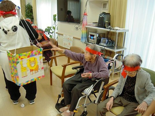 赤いハチマキをした高齢者たちが車いすやイスにしわって、パンだの格好をしたスタッフが箱を持っている。