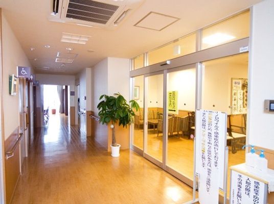 観葉植物が置かれた廊下は幅広く手すりが設置されている。硝子戸の向こうには応接セットが見える。