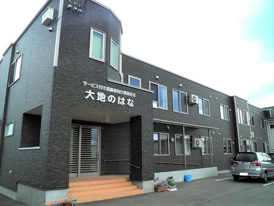 駐車場がついた２階建ての建物。玄関には階段と手すり付きのスロープ、花が植えられたプランターがある。