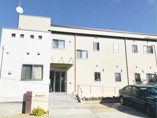 明るい雰囲気の２階建ての建物。玄関には手すり付きのスロープと階段があり、駐車場には車が止められ、花も植えられている。