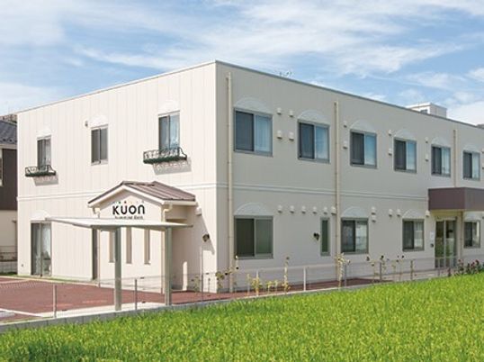 白を基調とした2階建ての建物。玄関先には屋根が付いていて、周辺には畑がある。