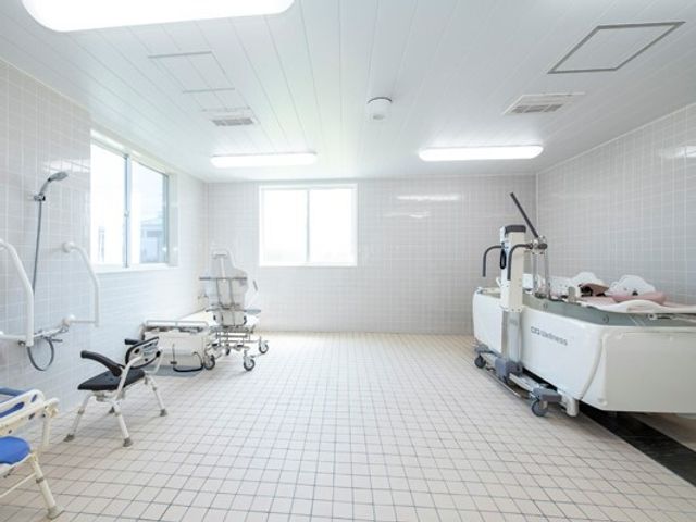 白を基調とした浴室はタイル張りで複数の窓が付き明るい雰囲気。右側には寝台浴が設置され、左側にはシャワースペースと座ったまま入浴できる機械浴がある。 ご入居者様の状態に応じた適切なサポートができるよう介護設備を充実させた住まい！お体の状態に合わせて福祉用具のレンタルも可能です。