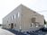 ベージュのレンガ調の外壁が印象的な2階建ての建物。2階部分にはベランダが付き、駐車スペースも用意され、周りには緑や住宅がある。
