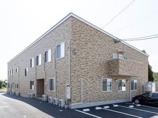 ベージュのレンガ調の外壁が印象的な2階建ての建物。2階部分にはベランダが付き、駐車スペースも用意され、周りには緑や住宅がある。