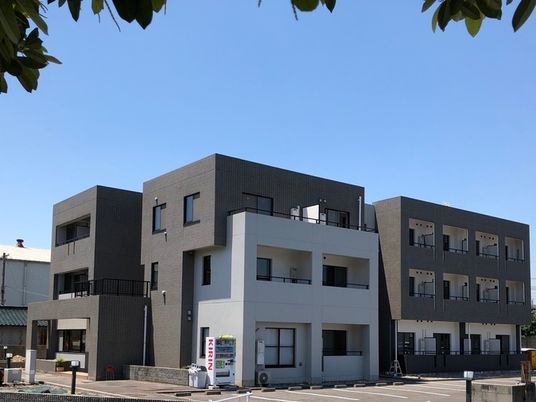 白とダークブラウンの３階建ての建物の前には自動販売機があり、日当たりのいい場所に建っている。