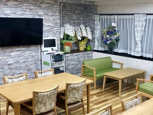 テレビが付いた食堂にはダイニングテーブルが複数置かれ、お祝いの花が飾られ、ソファセットもある。