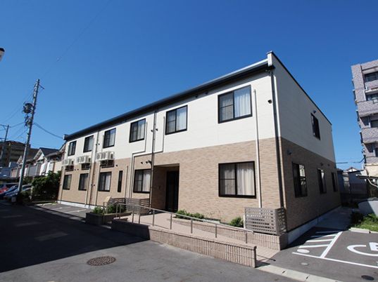 駐車スペースがあり、玄関には手すり付きのスロープが付いた2階建ての建物。外壁はフロアにより色が異なる。
