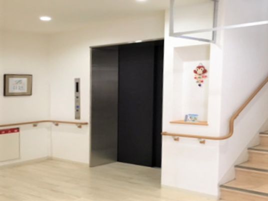 階段とエレベーターが並んで設置され、白い壁には木製の手すりが設置されている。大小の絵画も飾られ、清潔な空間。