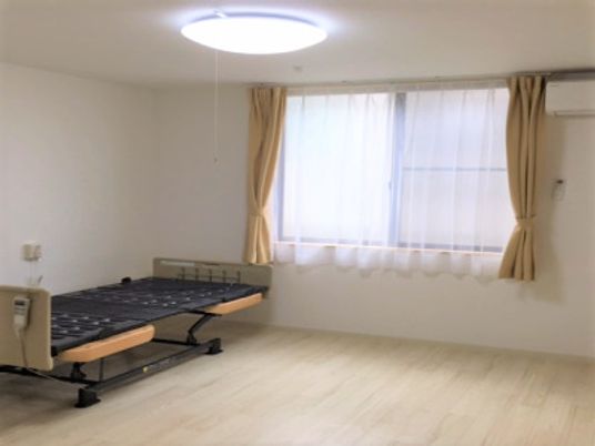 エアコンが付いたフローリングの部屋にはマットレスがない介護ベッドが置かれ、レースカーテンとカーテンが付いた大きな窓がある。
