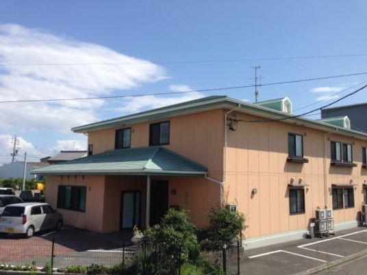 ベージュ系の外壁の２階建ての建物は日当たり良好で建物の前には車が止まっている。駐車場もある。