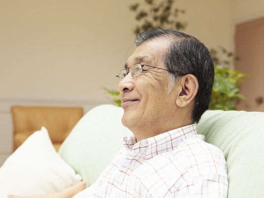 メガネをかけた高齢の男性がソファーに座りながら微笑みかけている様子。