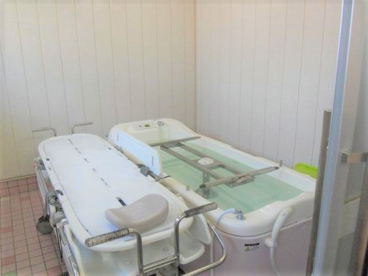 寝台浴が設置されたタイル張りの浴室。右側にはお湯が張られた浴槽型の機械が置かれ、左側にはストレッチャータイプのリフトが置かれている。