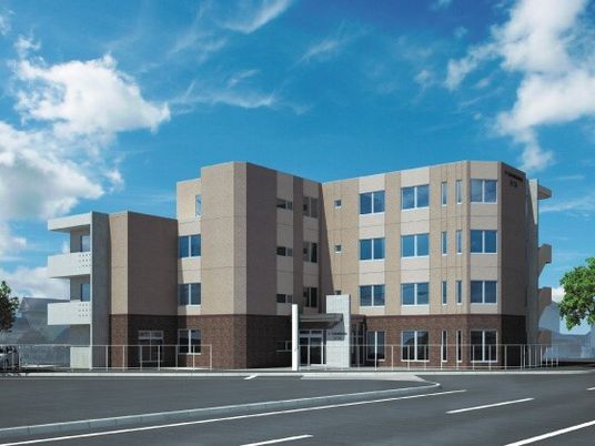 ベージュとブラウンの外壁の４階建ての建物の前には広い駐車場があり、玄関先には屋根が付いている。