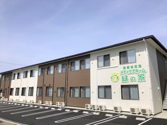 2階建ての建物はブラウンと白で落ち着いた雰囲気。玄関前にはスロープや緑がある。