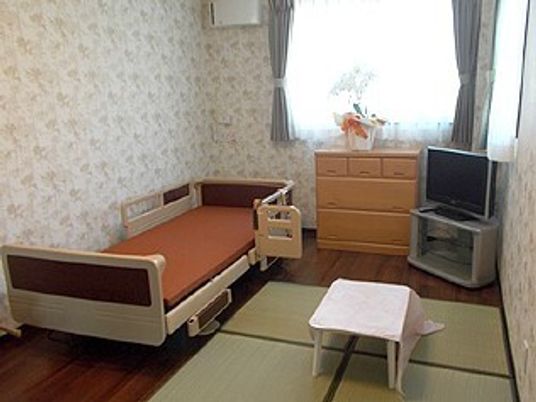 介護ベッドやテレビ、タンス、畳が置かれたフローリングの部屋には大きな窓が付き、カーテンが設置されている。タンスの上には花も飾られている。