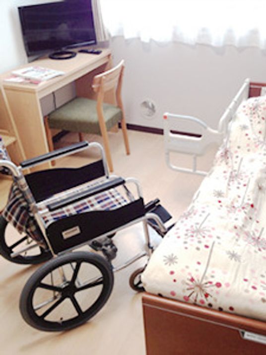 車いすが置かれたフローリングの部屋には寝具付きのベッドやテレビ、デスクセットが置かれている。