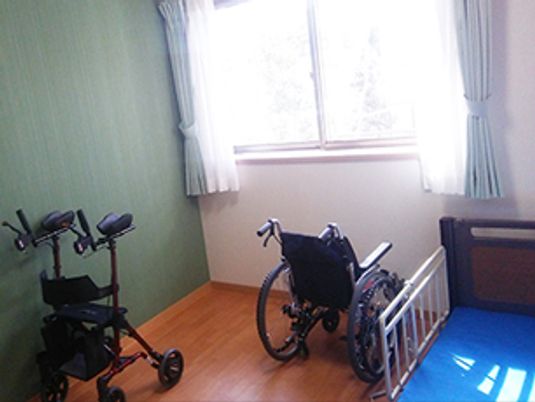 大きな窓があるフローリングの部屋には、車いすや歩行器、介護ベッドが置かれている。