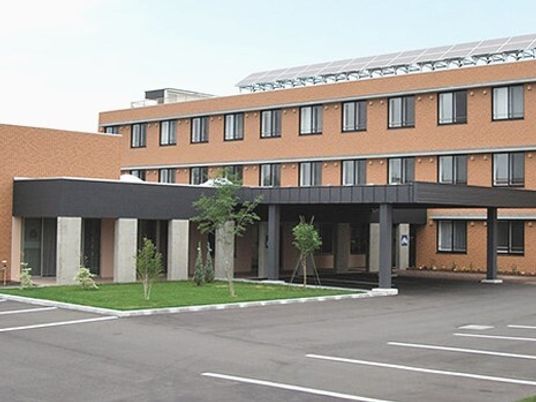 ブラウン系の３階建ての建物の玄関には車寄せが付き、芝生が植えられたスペースには草木も植えられている。
