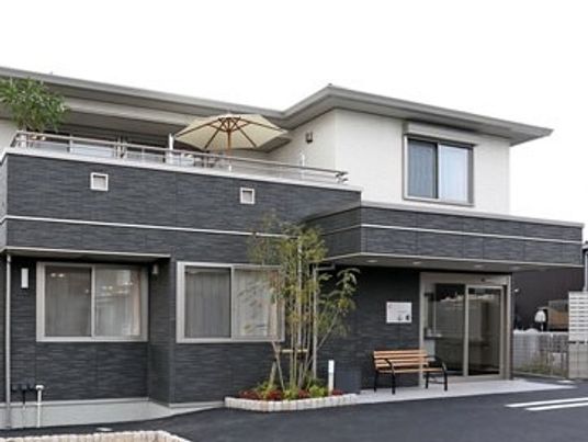 ダークグレーと白の２階建ての建物は玄関に屋根があり、木製のベンチが置かれ、駐車場も付いている。