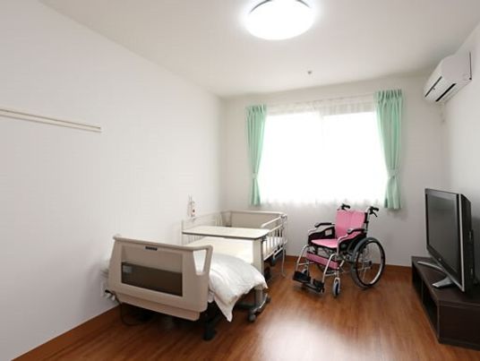 フローリングの部屋にはベッドやテレビ、車いす、エアコンがあり、緊急通報装置も付いている。