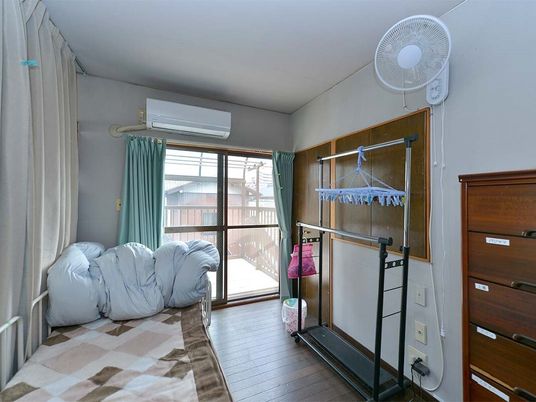 エアコンや扇風機が設置されたフローリングの部屋。寝具付きのベッドやタンス、ハンガーラックが置かれている。