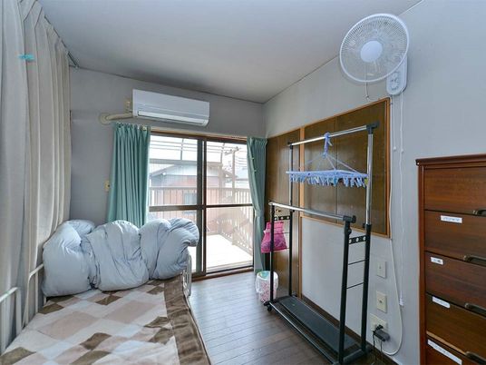 エアコンや扇風機が設置されたフローリングの部屋。寝具付きのベッドやタンス、ハンガーラックが置かれている。