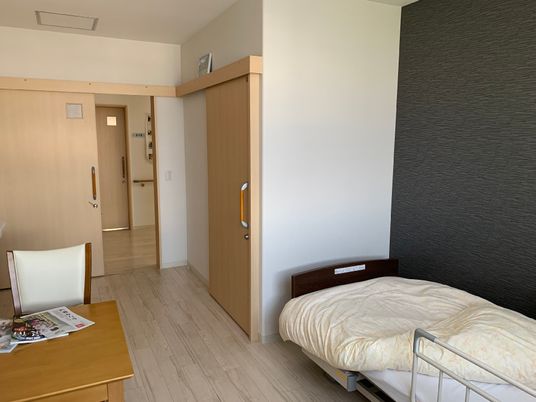 ベッドとテーブル、いすなどが置いてある施設内の居室。奥のほうに出入り口とトイレの入り口