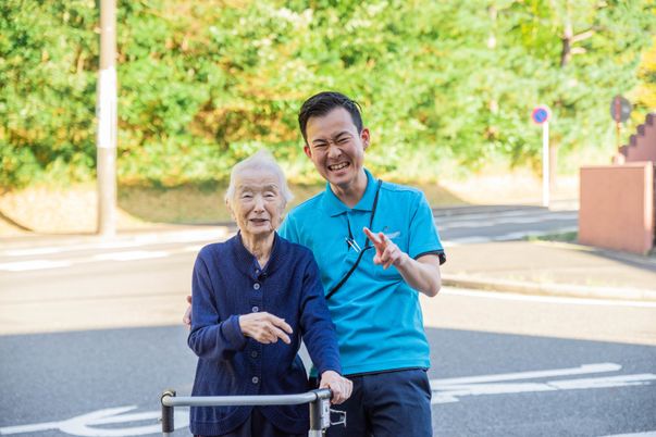 歩行補助器具をもってこちらを向いて微笑む高齢者と施設スタッフの様子