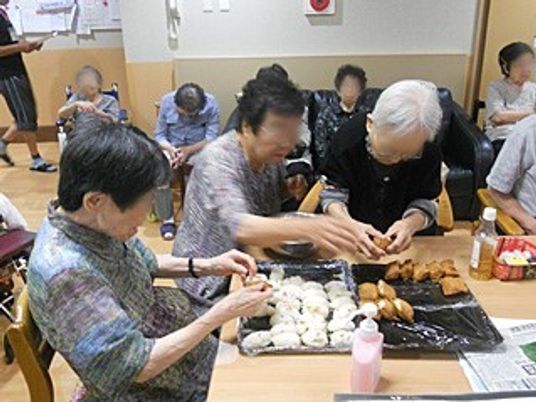 ３名の女性入居者がテーブルを囲んでいなり寿司を作っている様子。テーブルの上の黒いトレーには揚げを巻く前の寿司と、完成した寿司が並んでいる。