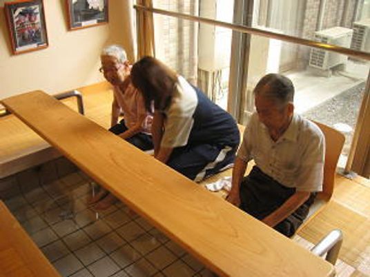 広い足湯に浸かっている男性入居者と女性入居者。二人の間にはジャージを着たスタッフが腰かけている。