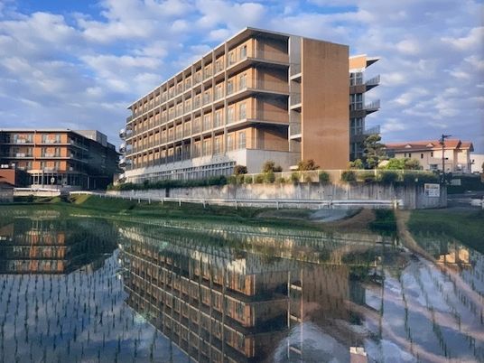 グレーの2階建ての建物の周りには緑が多く、建物の前には緑地スペースを中央に置いたロータリーもある。