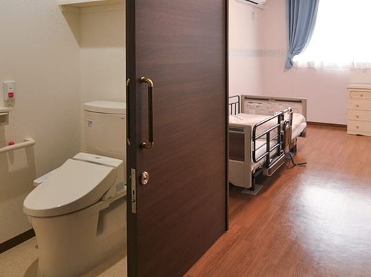 引き戸が付いたトイレが設置されたフローリングの居室にはタンスと介護ベッドが置かれている。