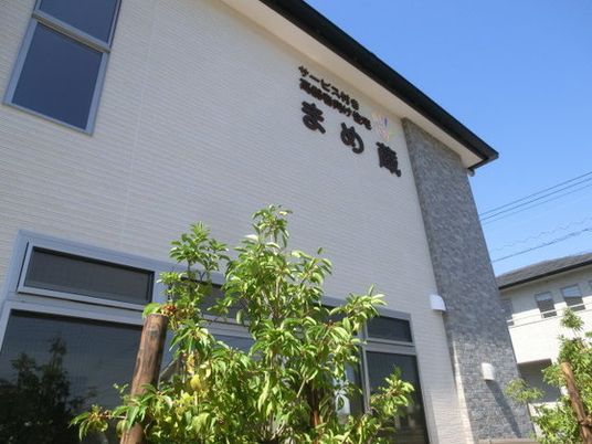 外壁に施設の名前が書かれた2階建ての建物。シンプルな色使いで周辺には緑が植えられている。