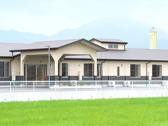 目の前に田んぼが広がっている平屋建ての建物。建物の周辺には白いフェンスがある。