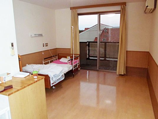 大きな窓が付いたフローリングの居室には寝具付きのベッドが設置され、エアコンが付き、窓にはカーテンがある。