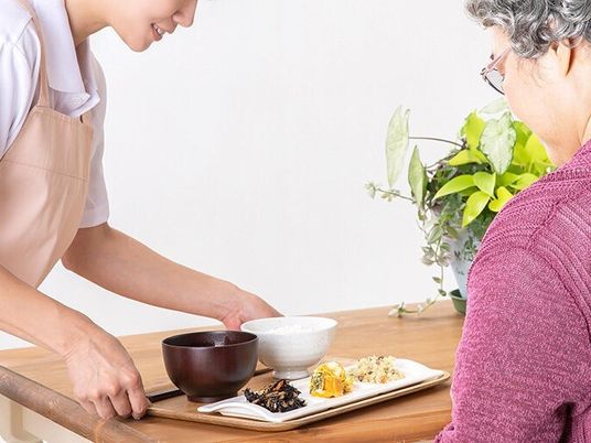 エプロンを付けた若い女性スタッフが高齢女性にトレーに乗った食事を提供している。