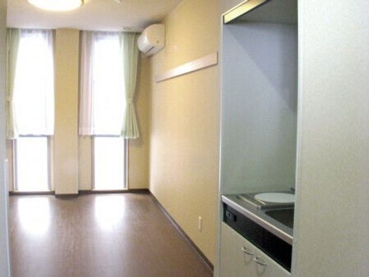 IHクッキングヒーター付きのキッチンが付いたフローリングの部屋にはエアコンがあり、カーテン付きの窓もある。