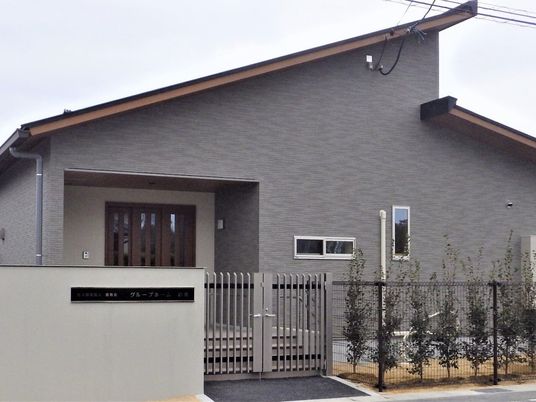 グレーの平屋建ての建物の周りには草木が植えられていて、フェンスが建てられ、大きな玄関のドアがある。